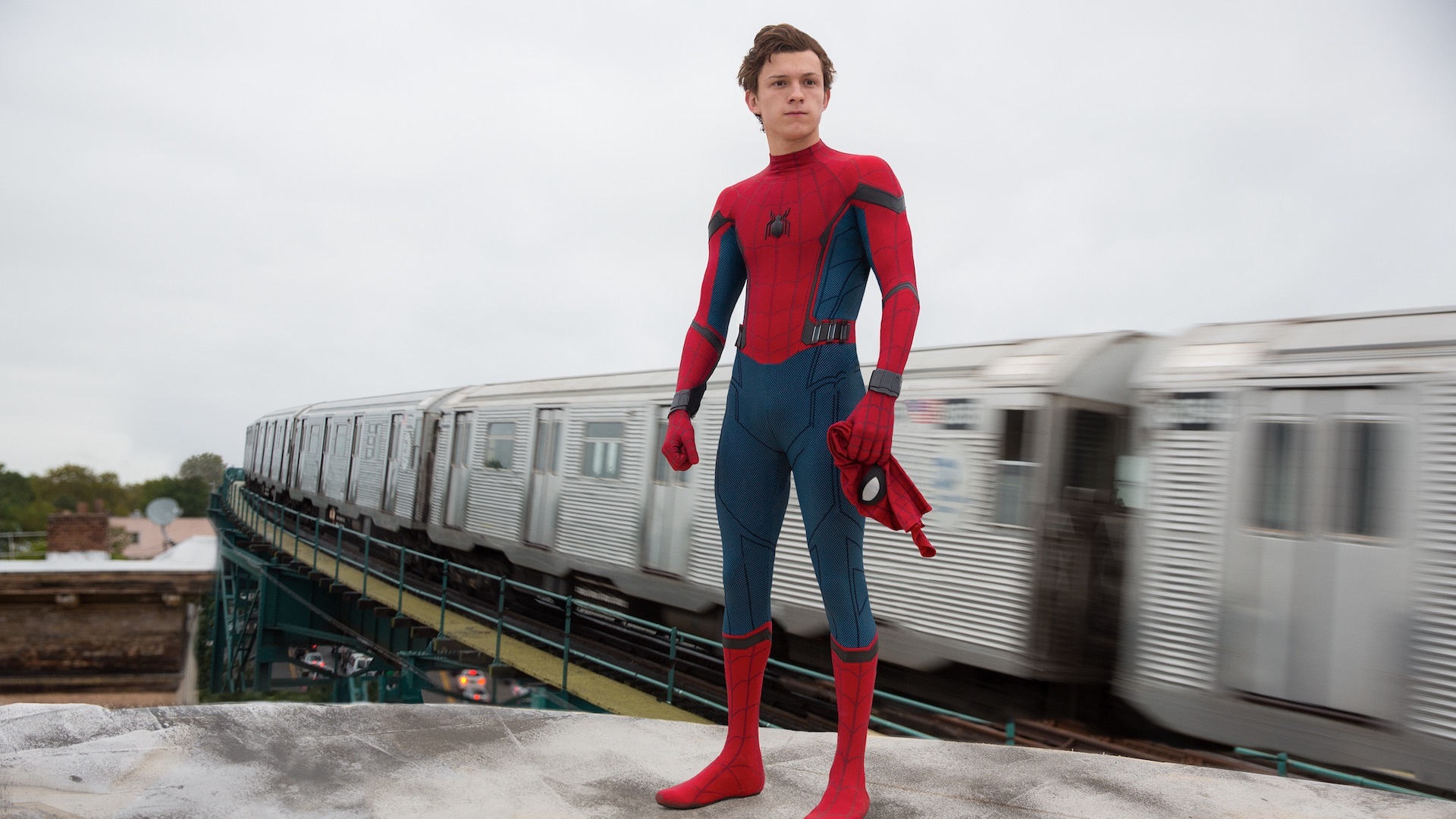 Tom Holland nei panni di Peter Parker, in piedi su un tetto e indossando l'abito di Spider-Man, ad eccezione della maschera che tiene in mano. Un treno della metropolitana di New York sfrenato sfreccia sullo sfondo.