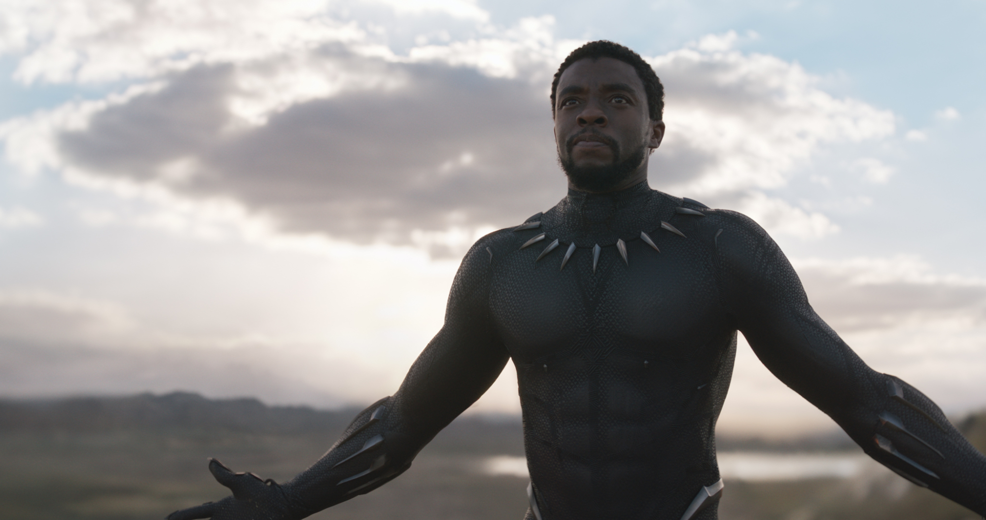 Un uomo (Chadwick Boseman nei panni di T'Challa) in un elegante abito nero da supereroe cammina con sicurezza con le braccia aperte, l'orizzonte dietro di lui.