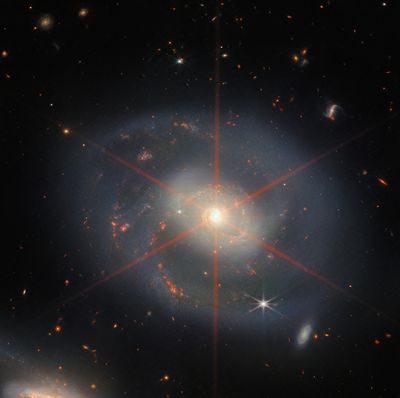 Questa immagine mostra una galassia a spirale dominata da una regione centrale luminosa. La galassia ha sfumature blu-viola con regioni rosso-arancio piene di stelle. È visibile anche un grande picco di diffrazione, che appare come uno schema a stella sulla regione centrale della galassia. Molte stelle e galassie riempiono la scena sullo sfondo